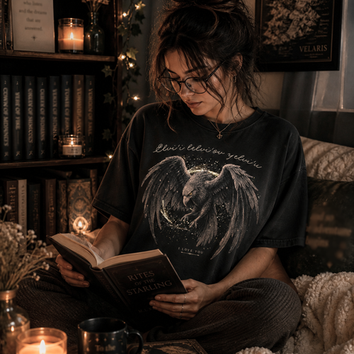 Woman wearing a romantasy graphic tee with a winged bird design and script text, sitting in a cozy candlelit reading space with books and warm lighting